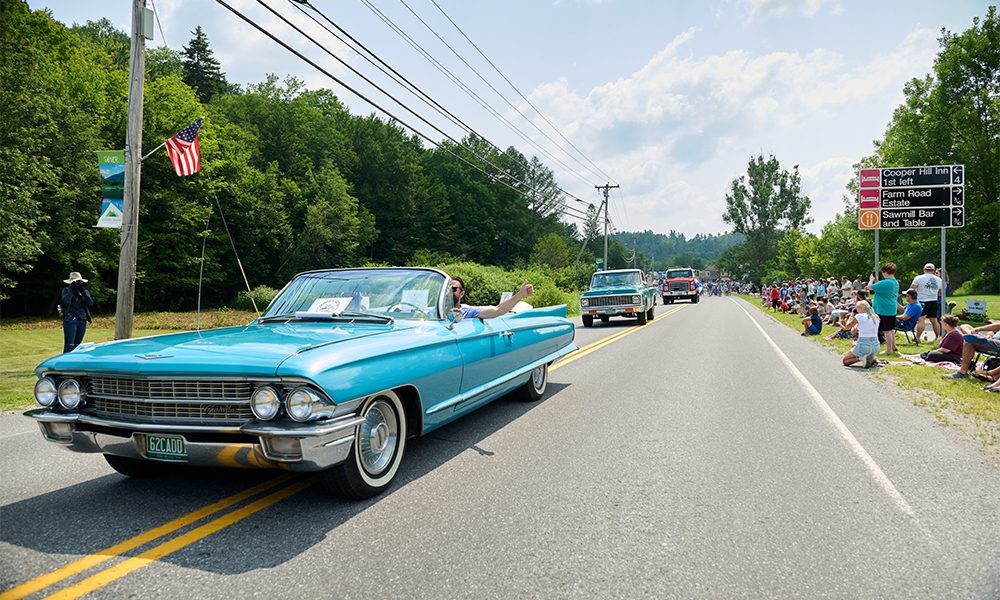A turquoise classic convertible car leads a parade down a rural road, with people sitting on both sides and American flags visible in the background.