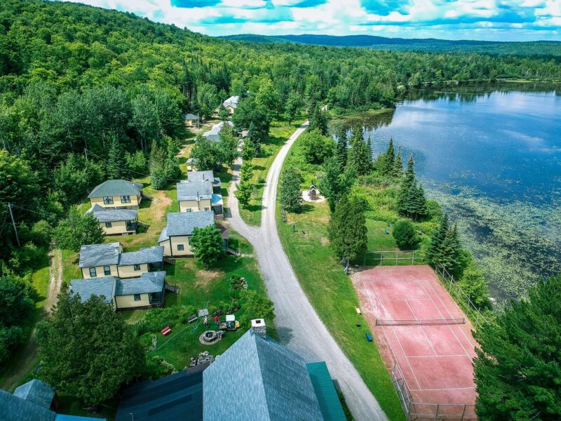 Aerial view of lakeside cabins, a tree-lined road, tennis court, and forested landscape under a partly cloudy sky.