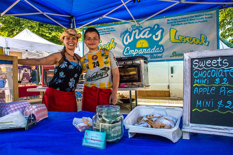 Woman and boy smile at a farmers market empanada stall under a blue canopy. Chalkboard lists sweets and mini empanadas.