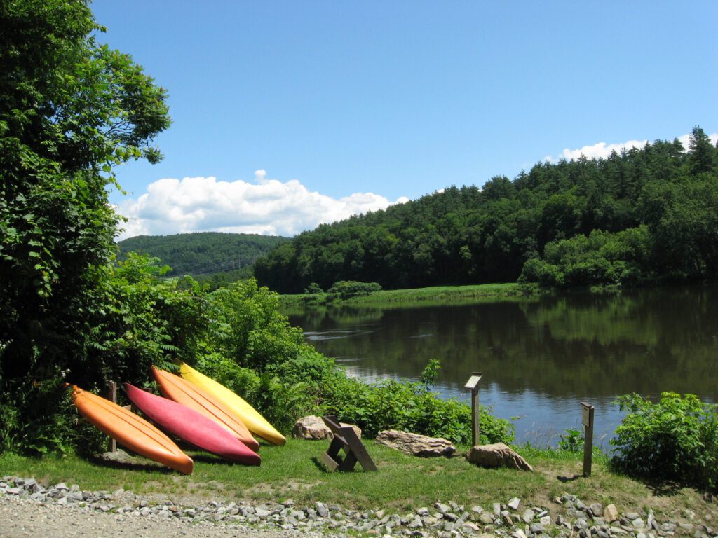 Four colorful kayaks rest on grassy ground beside a calm river, with lush green trees and hills in the background under a blue sky with clouds.
