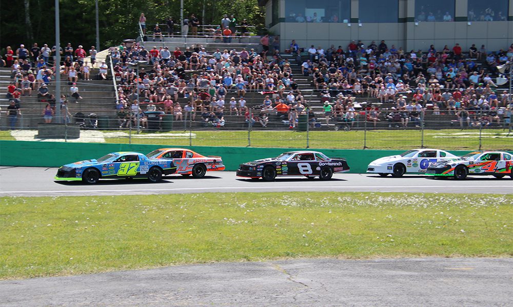 Race cars drive around a track under a clear sky, cheered on by a large crowd in the stands.
