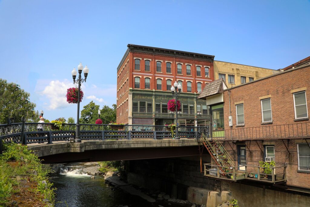 A historic brick building sits beside a bridge adorned with hanging flower baskets and street lamps. A river flows beneath, under a clear blue sky.