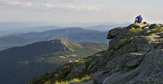 A person sitting on a high mountain peak with other mountains visible in the background. 