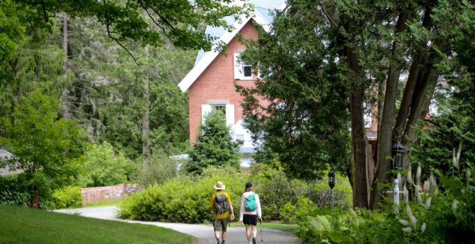 Two hikers stroll down a gravel path surrounded by lush greenery, approaching a charming red brick house.