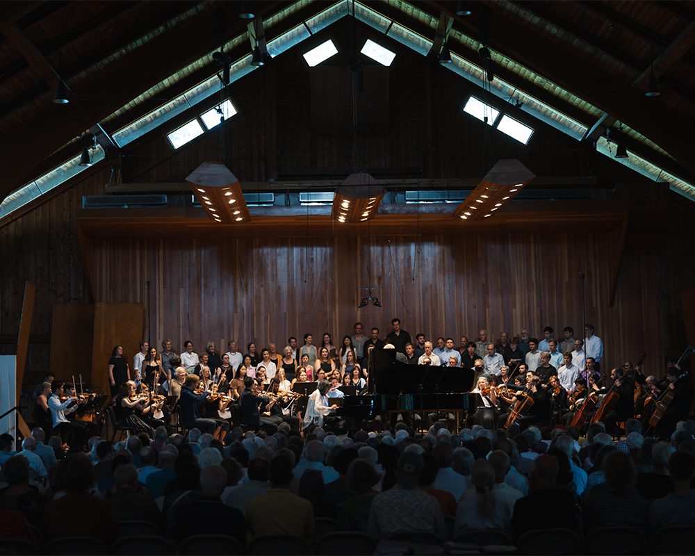 A full orchestra and choir perform on stage in a wooden concert hall with a large audience seated in front.