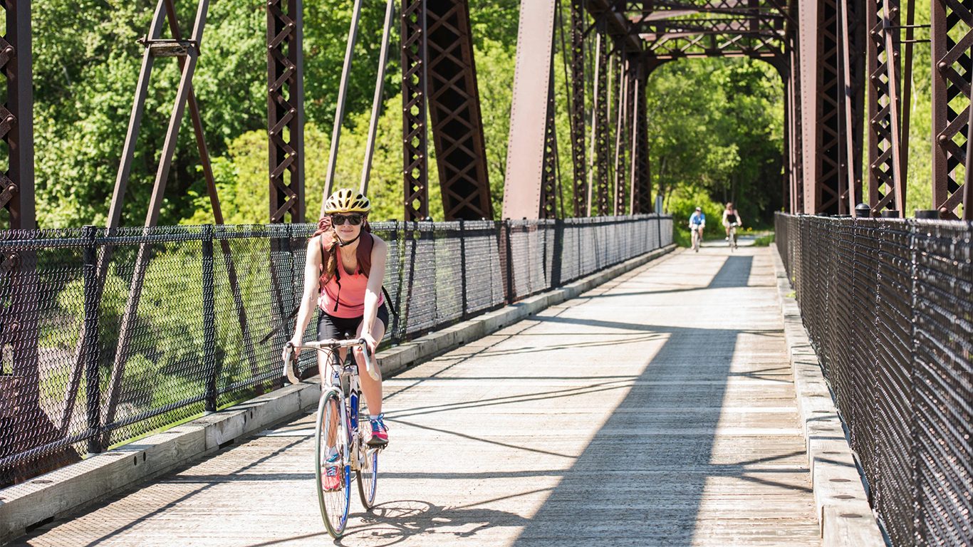 A person wearing a helmet rides a bicycle across a metal bridge on a sunny day, with two other cyclists in the background.