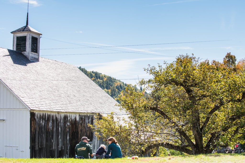 Two adults and a child in front of a wooden barn and a tree on a spring day with the sun out and a clear blue sky.