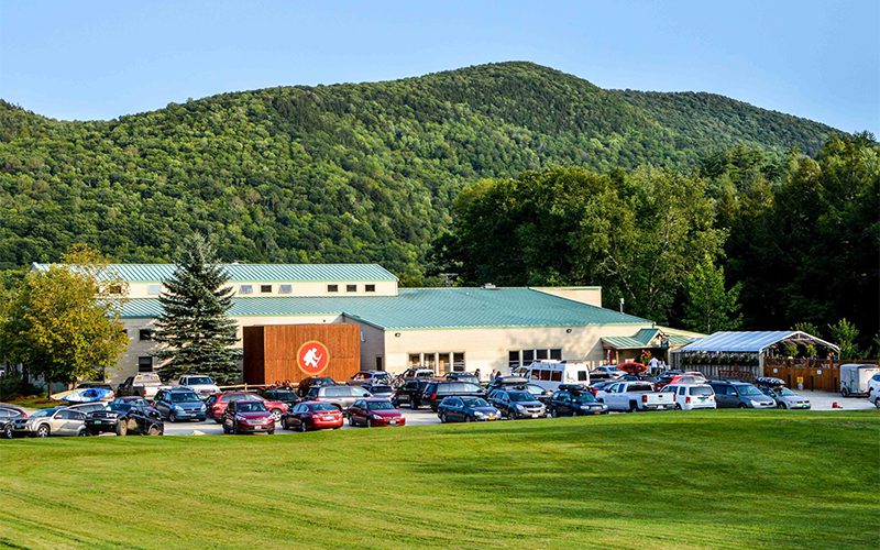 A spacious building with a green roof, surrounded by trees and mountains, filled with parked cars in a sunny, outdoor setting.