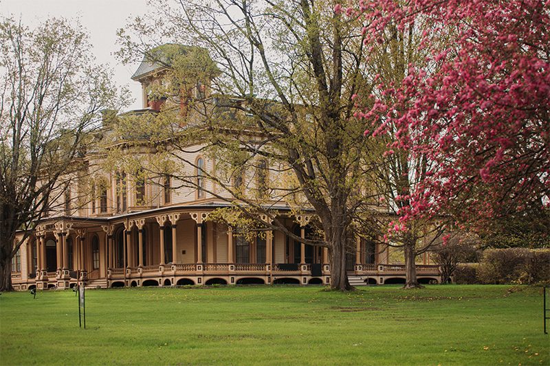 A historic mansion with ornate details, surrounded by trees and blooming pink flowers, set on a lush green lawn.