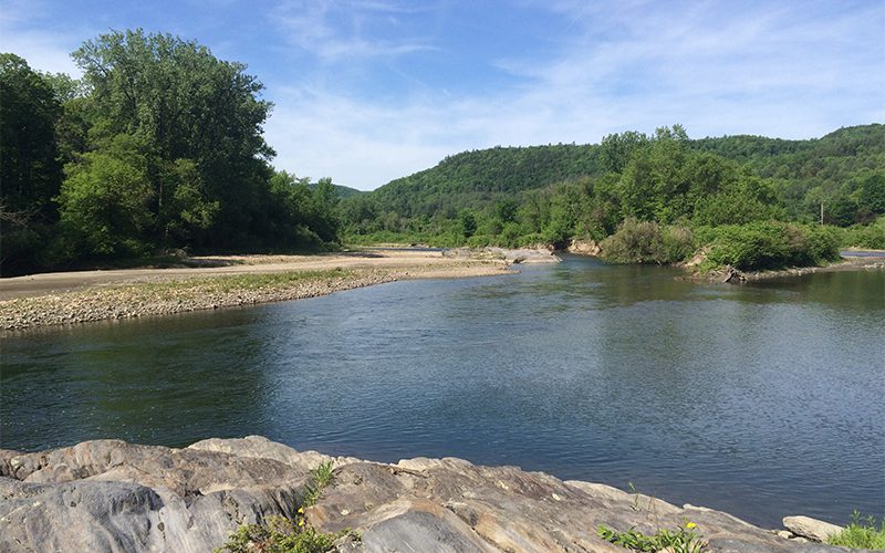 A serene river curves through lush green landscapes, flanked by hills under a clear blue sky, with rocky terrain in the foreground.
