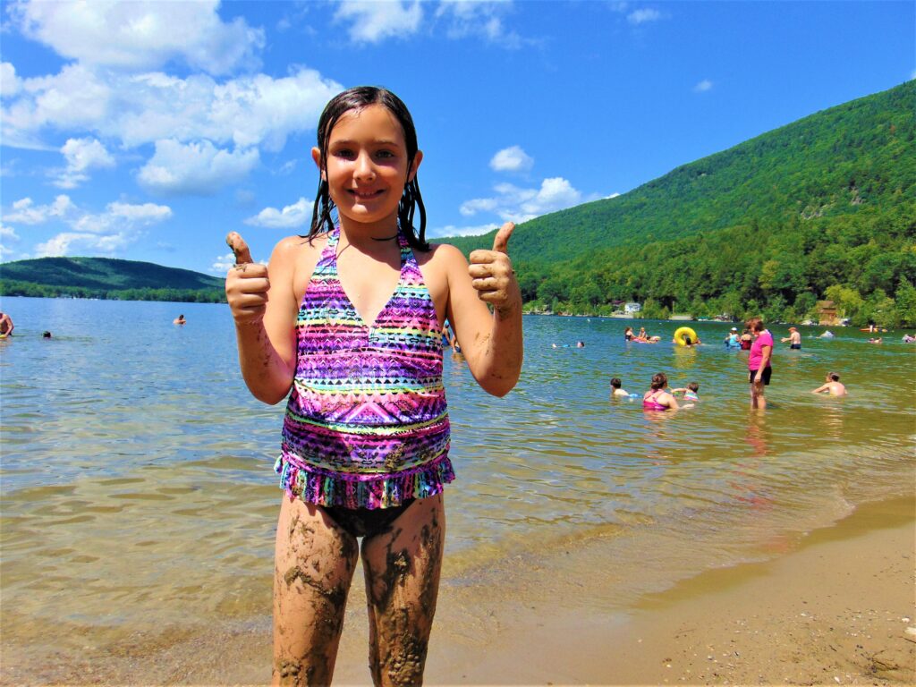 A smiling girl in a colorful swimsuit gives thumbs up on a sandy lakeside. Her legs are muddy, with people swimming nearby under a clear blue sky.