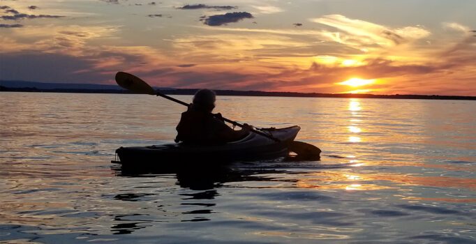 A person kayaks on calm water during a vibrant sunset, with colorful clouds reflecting on the surface, creating a peaceful and tranquil scene.