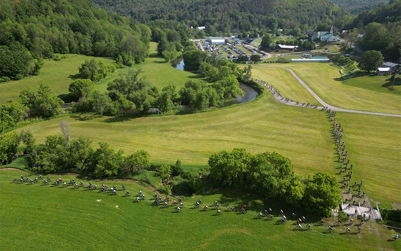Aerial view of a winding motorcycle convoy through lush green fields, with a small town and parking area in the background.