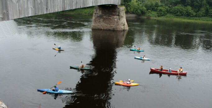 Several people paddle kayaks and a canoe on a river beneath a wooden bridge, with trees and greenery along the riverbank in the background.