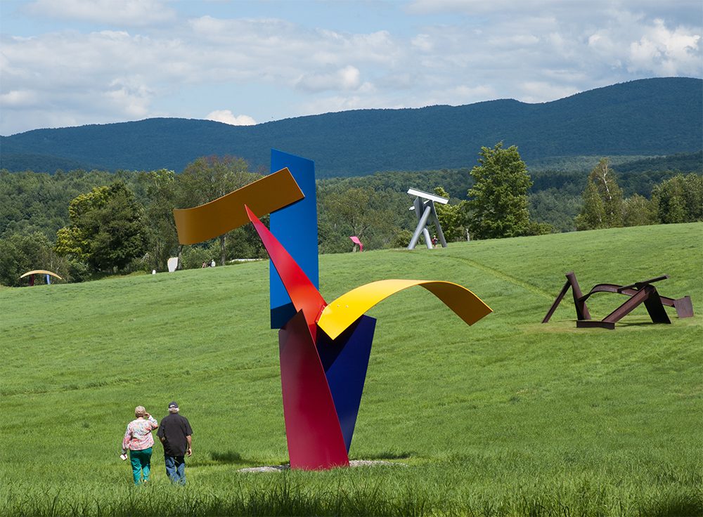 A vibrant abstract sculpture in red, blue, and yellow stands on a lush green field. Two people walk nearby, with mountains in the background under a partly cloudy sky.