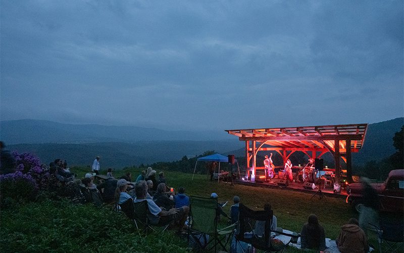 A live band performs on a scenic stage at dusk, with an audience seated on the lawn, surrounded by mountains and colorful flowers.