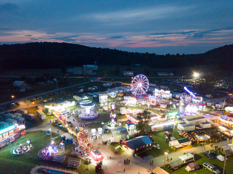 Aerial view of a lit-up fairground at dusk, featuring vibrant rides, a Ferris wheel, and bustling crowds, set against a serene pastel sky and dark hills.