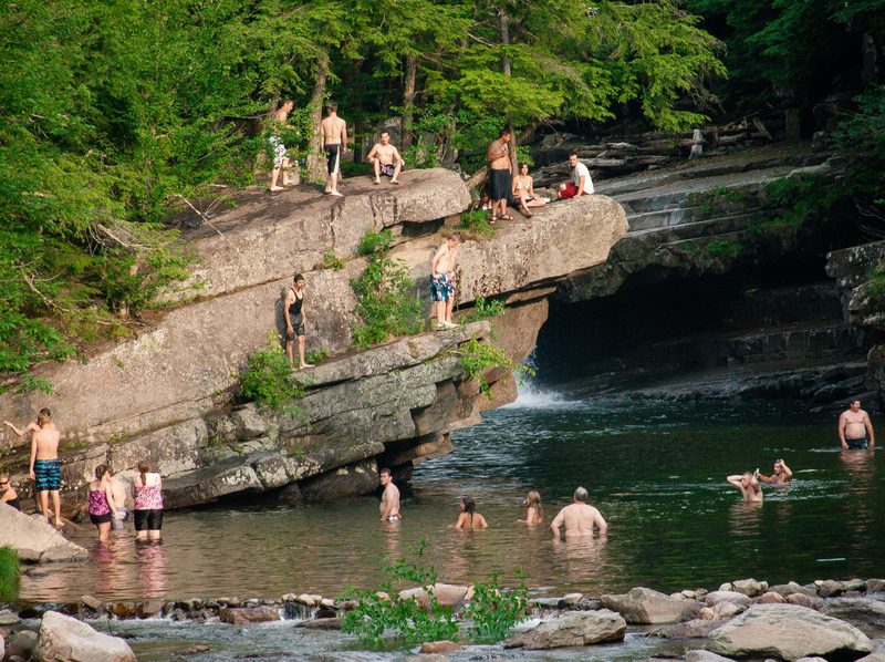 People enjoying a sunny day at a natural swimming hole surrounded by lush greenery. Some individuals stand on a rock ledge, while others swim below.