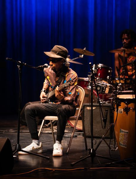 A musician performs on stage, seated with a guitar, wearing a colorful shirt and hat, against a vibrant blue backdrop.