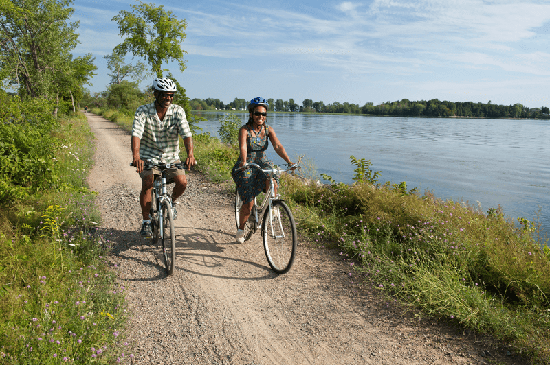 Two people riding bikes along a lakeside trail on a sunny day, surrounded by lush greenery and clear blue skies.