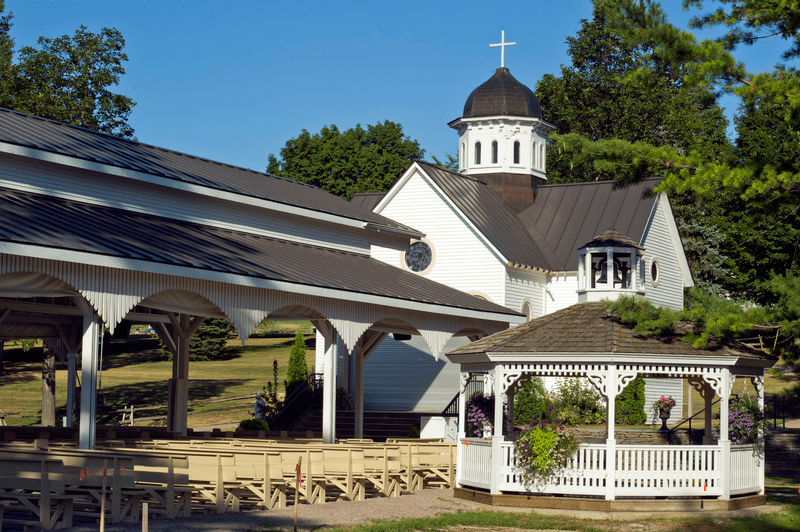 Outdoor church scene with a white gazebo, covered seating area, and a white building topped by a cross. Surrounded by lush green trees under a clear blue sky.