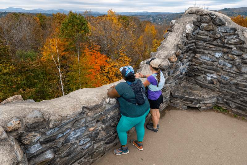 Two people on top of a stone tower admiring a view of fall foliage.