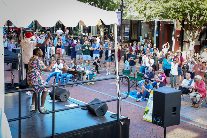 A group of people stand around an outdoor stage watching someone sing.