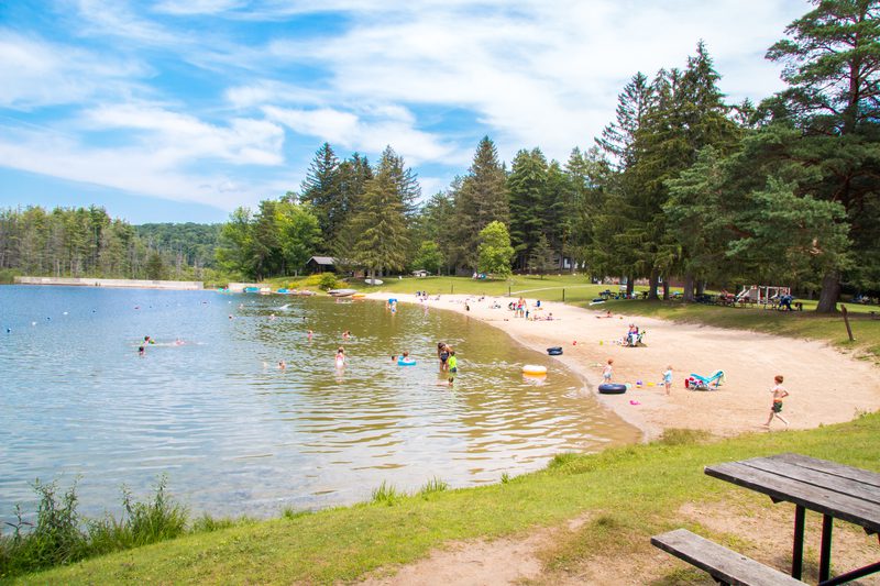 Seen from afar, people sit on a sandy beach by a lake. Some are swimming. It’s a sunny summer day.