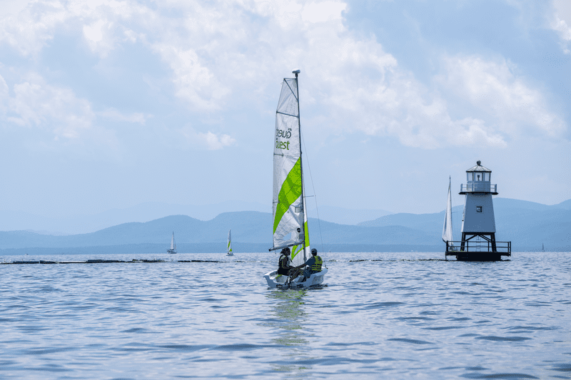 Sailboat with green and white sail glides on blue water under cloudy sky's near a small lighthouse. Mountains can be seen from a distance.