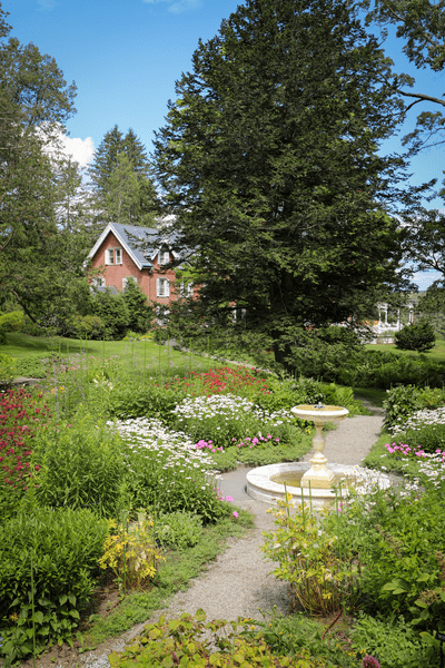 A lush garden with colorful flowers, a white fountain, and a charming red house in the background under a clear blue sky.