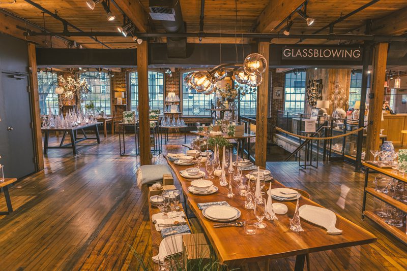 A spacious, rustic glass art studio featuring elegant glassware displays, a dining table set, and a sign for glassblowing workshops.