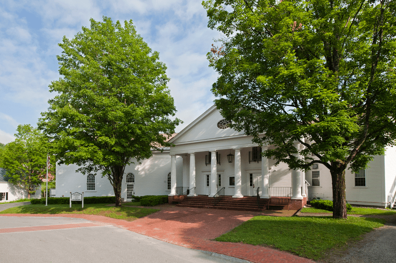 White building with classical columns, framed by lush green trees on a sunny day.