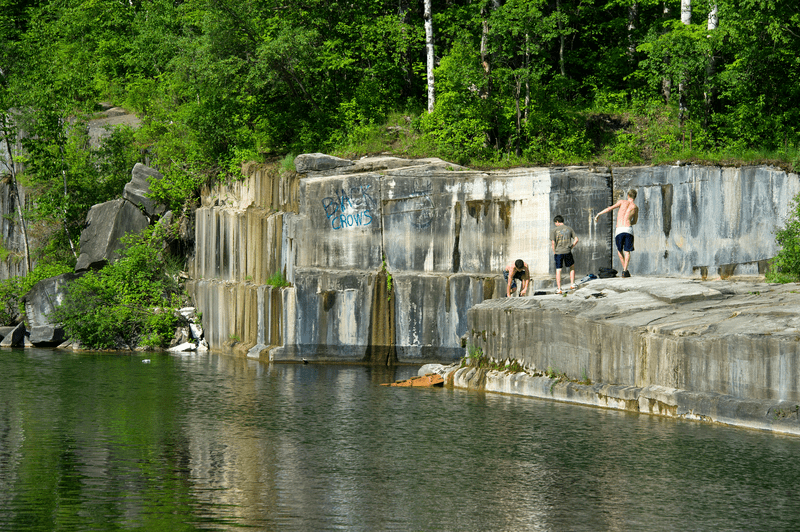 Three people play on a rocky shoreline, with lush greenery in the background and calm water reflecting the sunlight.