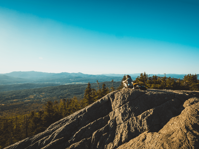A hiker sits atop a rocky outcrop, gazing at a vast, scenic landscape of mountains and forests under a clear blue sky.
