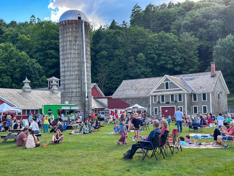 A lively outdoor gathering at a farm with a tall silo and rustic buildings. People are sitting on blankets and chairs, enjoying a sunny day amidst greenery.
