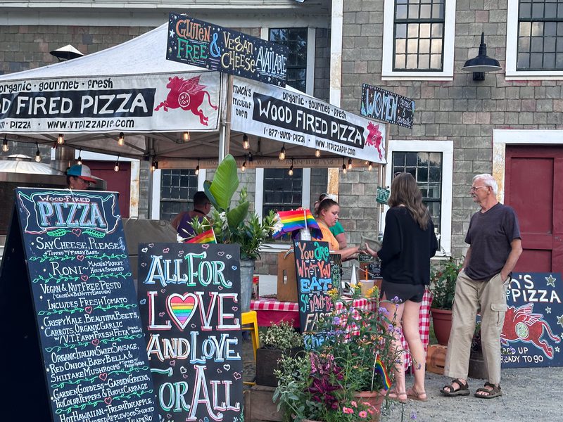 Outdoor wood-fired pizza stand with several colorful signs showing menu options and people talking to staff at a table.