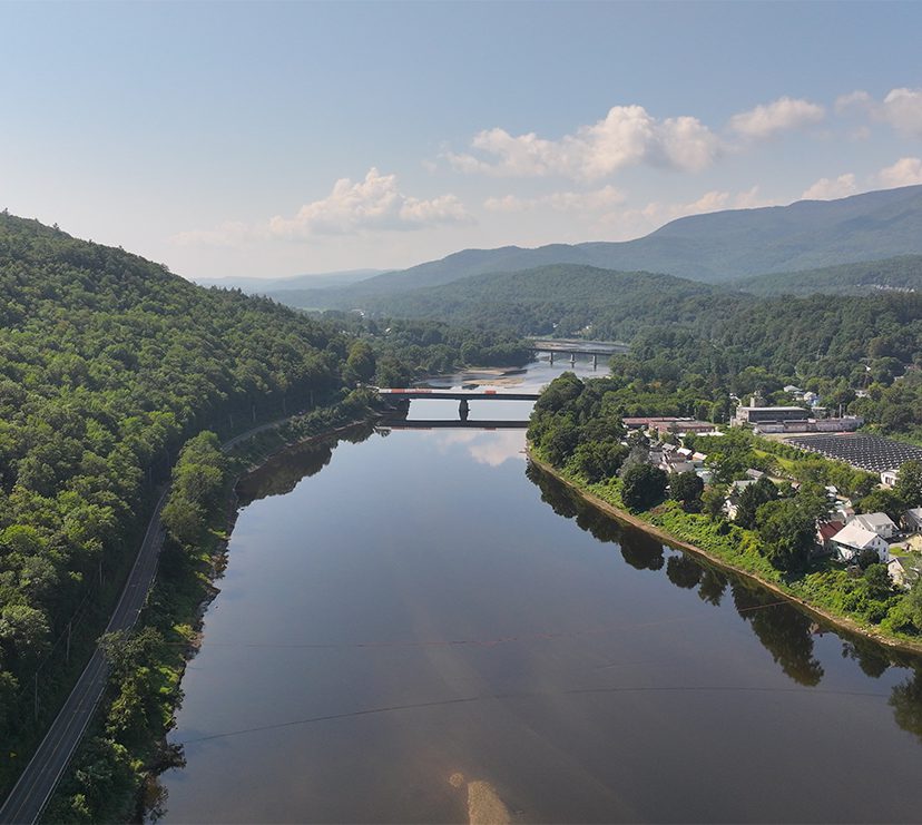 Seen from above, a bridge spans a river in the summer. Green trees and mountains are visible in the area surrounding a downtown.
