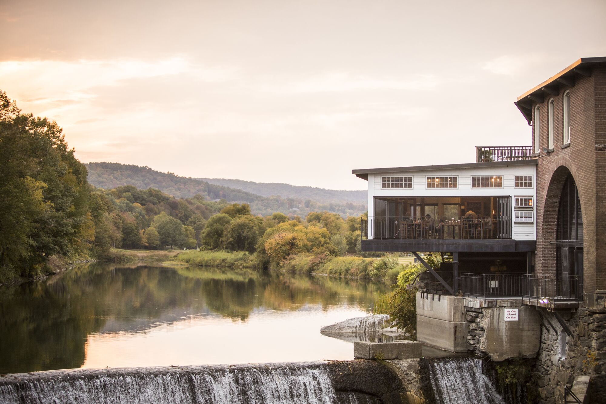 A restaurant that overlooks a river with a rushing waterfall seen from outside in the summer at sunset.