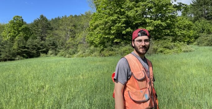 Andrew Hirsch, a young man in an orange forestry vest in a green meadow.