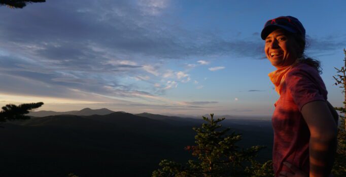 A person smiling with a mountain range behind them at dusk.