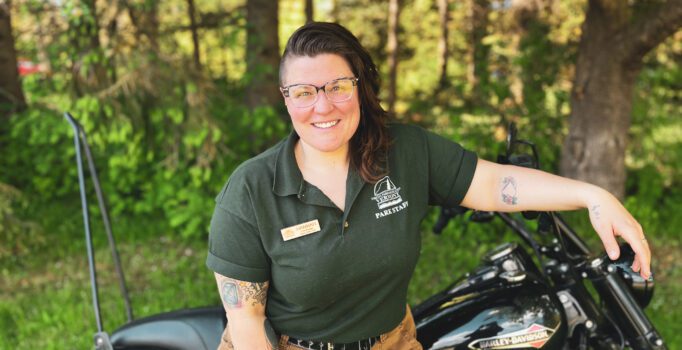A person in front of a motorcycle smiling at the camera outside in the summer. 