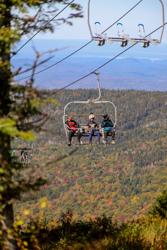 Three people on a chair lift surrounded by red, yellow and orange fall foliage.