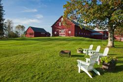 White Adirondack chairs and a fire pit sit on a spacious, sloping green lawn in front of a classic red barn and farmhouse under a partly cloudy blue sky.