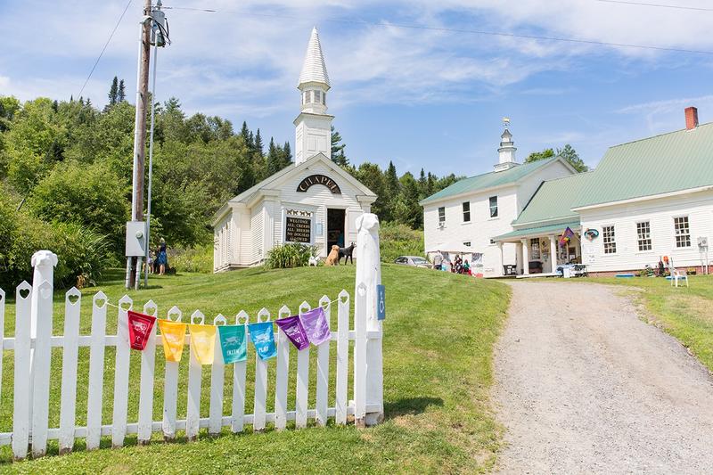 Colorful flags hang on a white picket fence outside a chapel in the summer. 