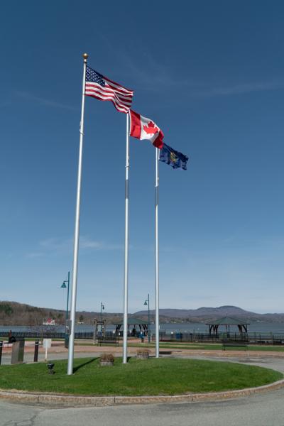 An American flag, a Canadian flag, and a Vermont State flag blows in the breeze as the lake and the mountains are in the background.