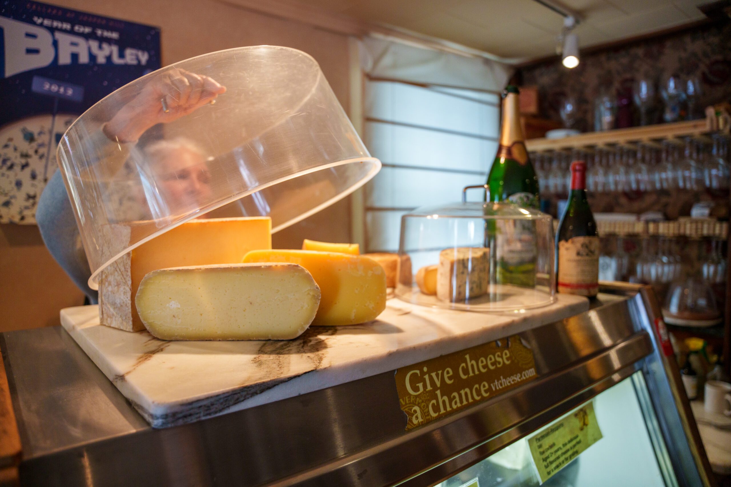 A cheese seller lifts a glass lid covering wheels of cheese in Vermont.