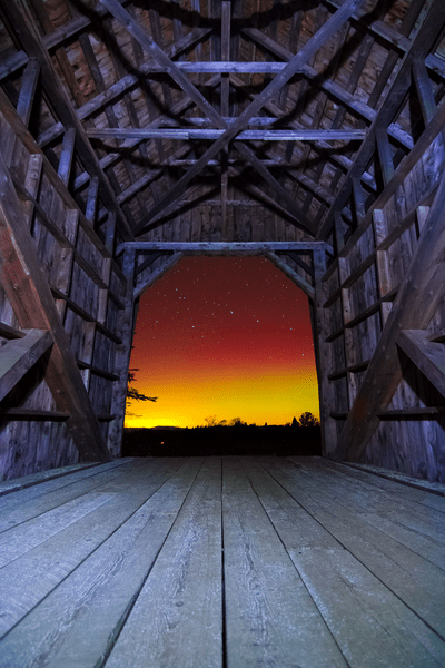The aurora borealis, in bright colors, visible through a covered bridge at night.