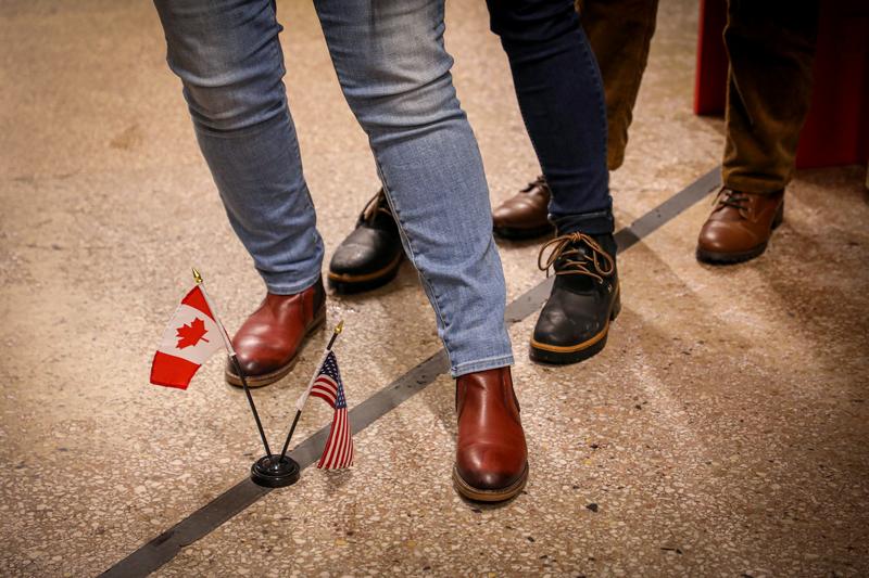 Three sets of feet standing on either side of the U.S.-Canada border.