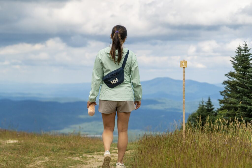 A hiker wearing a light jacket and a cross-body bag walks along a grassy mountain ridge overlooking distant blue hills under a cloudy sky.