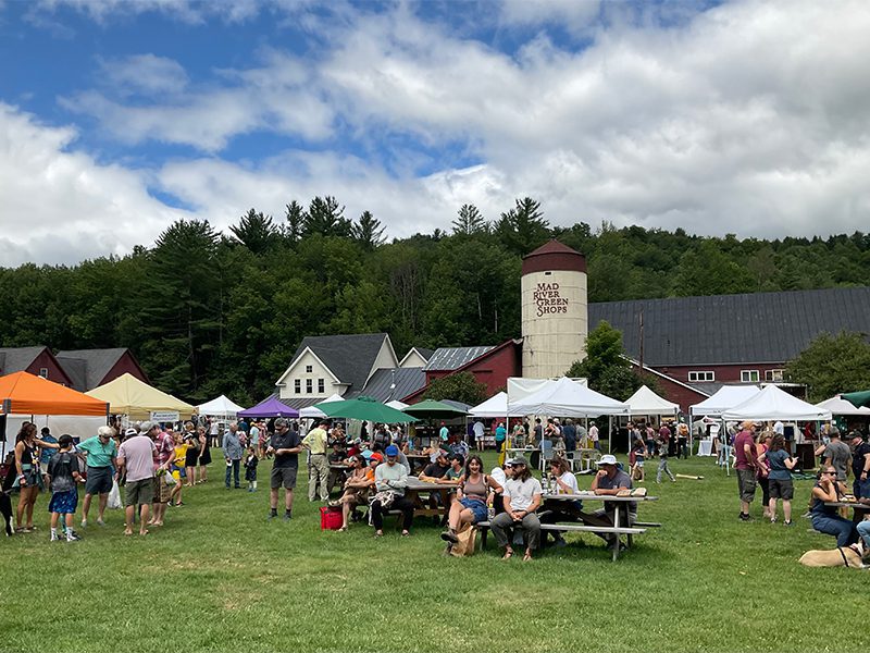 People browse at outdoor stalls at a market on a sunny summer day. A silo is behind them. 